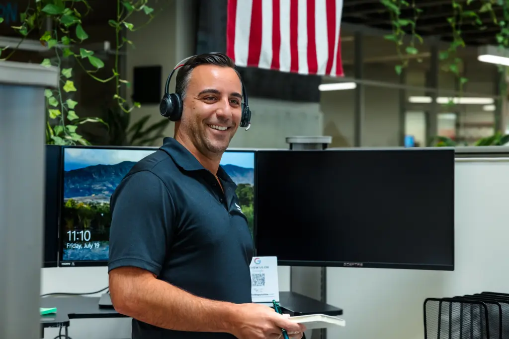 A man wearing a headset and a dark polo shirt smiles while standing in an office with computer monitors and an American flag in the background.