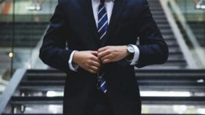 A person in a dark suit and striped tie buttons their jacket while standing in front of blurred stairs. Their face is not visible, and they are wearing a wristwatch.