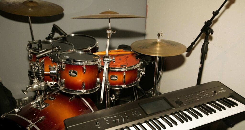 A drum set with brown sunburst finish sits near a digital keyboard and a microphone stand, all positioned in a music rehearsal or recording space.