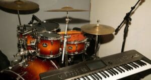 A drum set with brown sunburst finish sits near a digital keyboard and a microphone stand, all positioned in a music rehearsal or recording space.